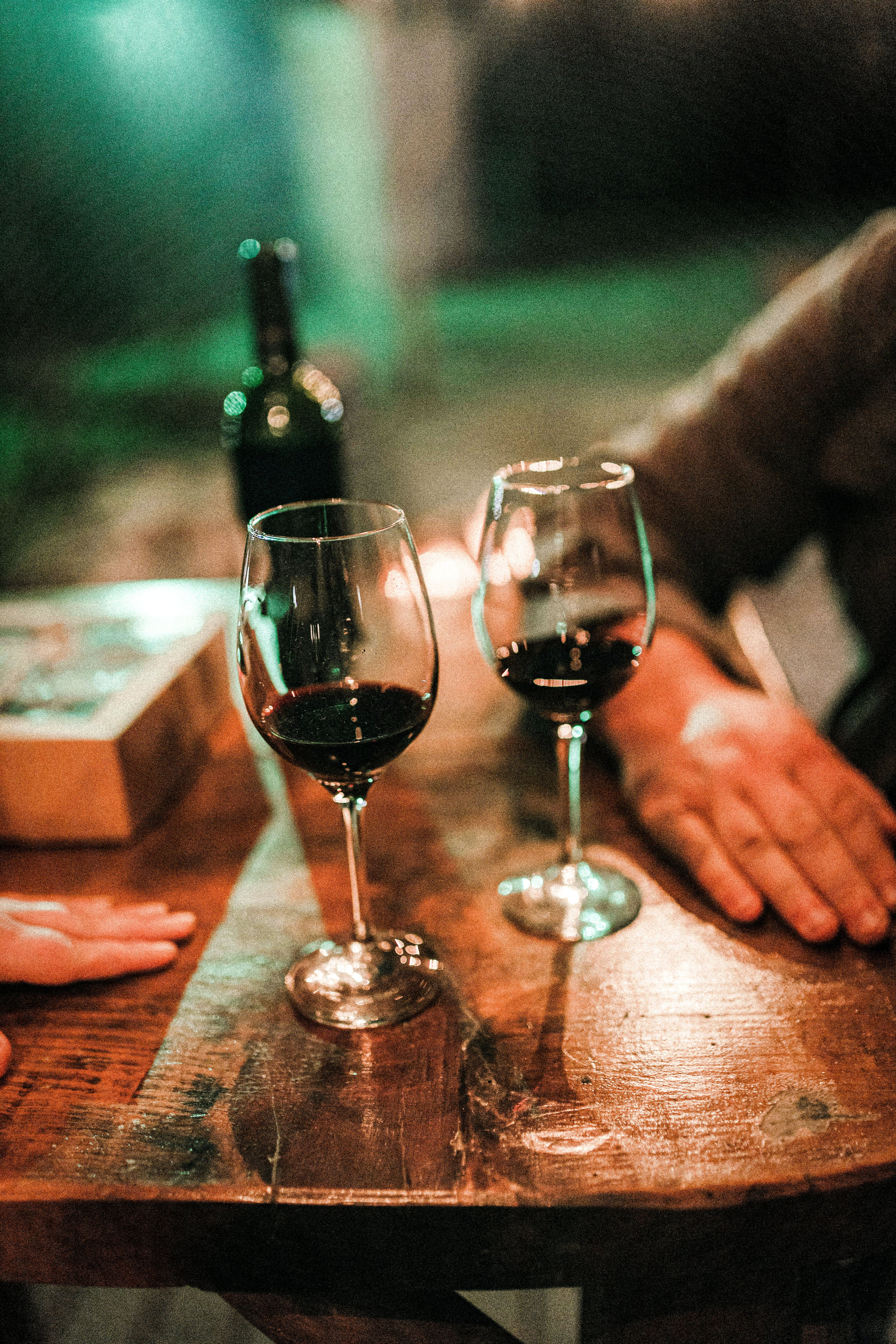 Two wine glasses with red wine on a wooden table, with a bottle and blurred background.