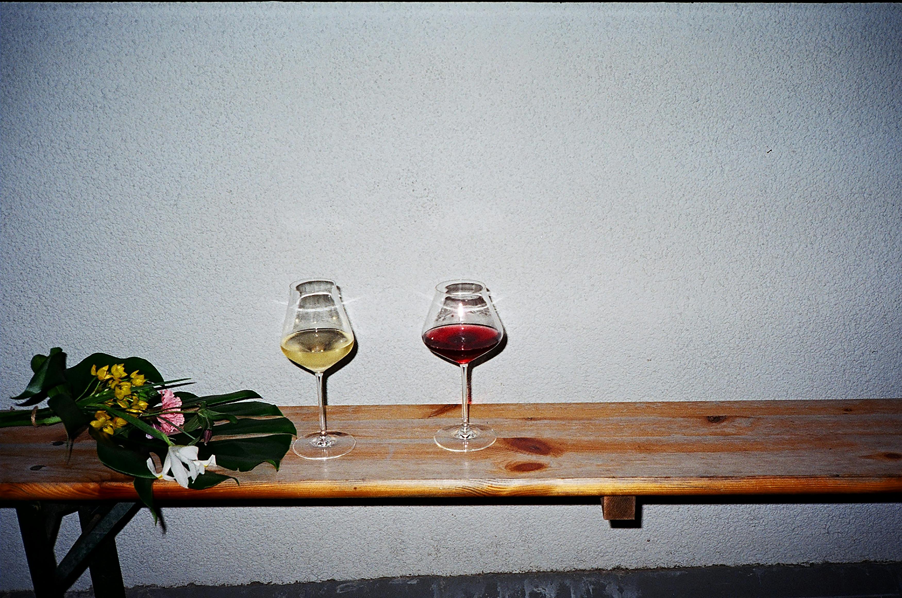 Two wine glasses with different colored liquids on a wooden surface against a plain wall.